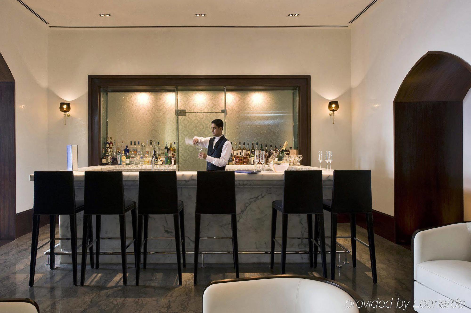 The Taj Mahal Tower, Mumbai Hotel Restaurant foto The photo shows a stylish bar area. In the center, a bartender, dressed in a formal outfit, is standing behind a sleek, modern bar counter made of marble. The bar is lined with high chairs for patrons. Behind the bartender, there are shelves filled w